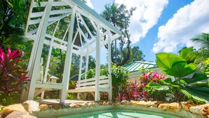 Un belvédère blanc avec une piscine en pierre, entouré de plantes tropicales et surplombé d’un ciel d’un bleu limpide.