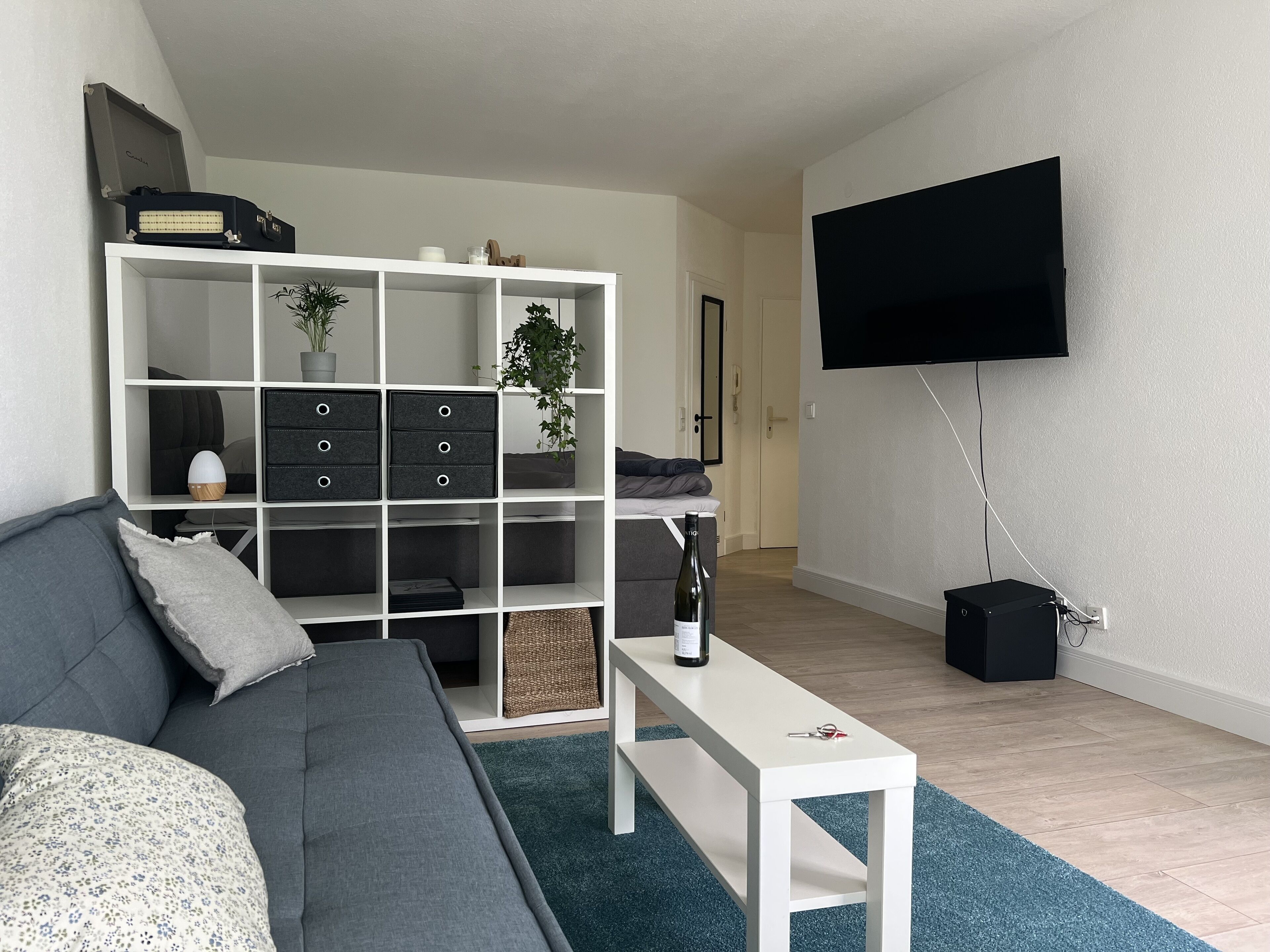 Ein modernes Wohnzimmer mit grauem Sofa, einem weißen Couchtisch, einer weißen Regaleinheit mit Stauraum, einem flachbildfernseher an der Wand und einem kleinen schwarzen Lautsprecher.