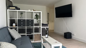 A modern living room with a grey sofa, a white coffee table, a white shelving unit with storage, a flat-screen TV mounted on the wall, and a small black speaker. - Wunderschöne 1-zimmer Wohnung (Wiesbaden)