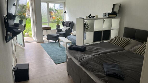 A modern bedroom with a large bed, a sofa, a desk, and a chair. There is a sliding door leading to a balcony with plants and a view of the outside. - Wunderschöne 1-zimmer Wohnung (Wiesbaden)
