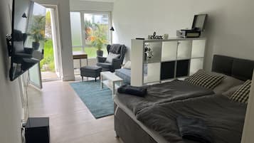 Ein modernes Schlafzimmer mit einem großen Bett, einem Sofa, einem Schreibtisch und einem Sessel. Eine Schiebetür führt zu einem Balkon mit Pflanzen und Blick ins Freie.