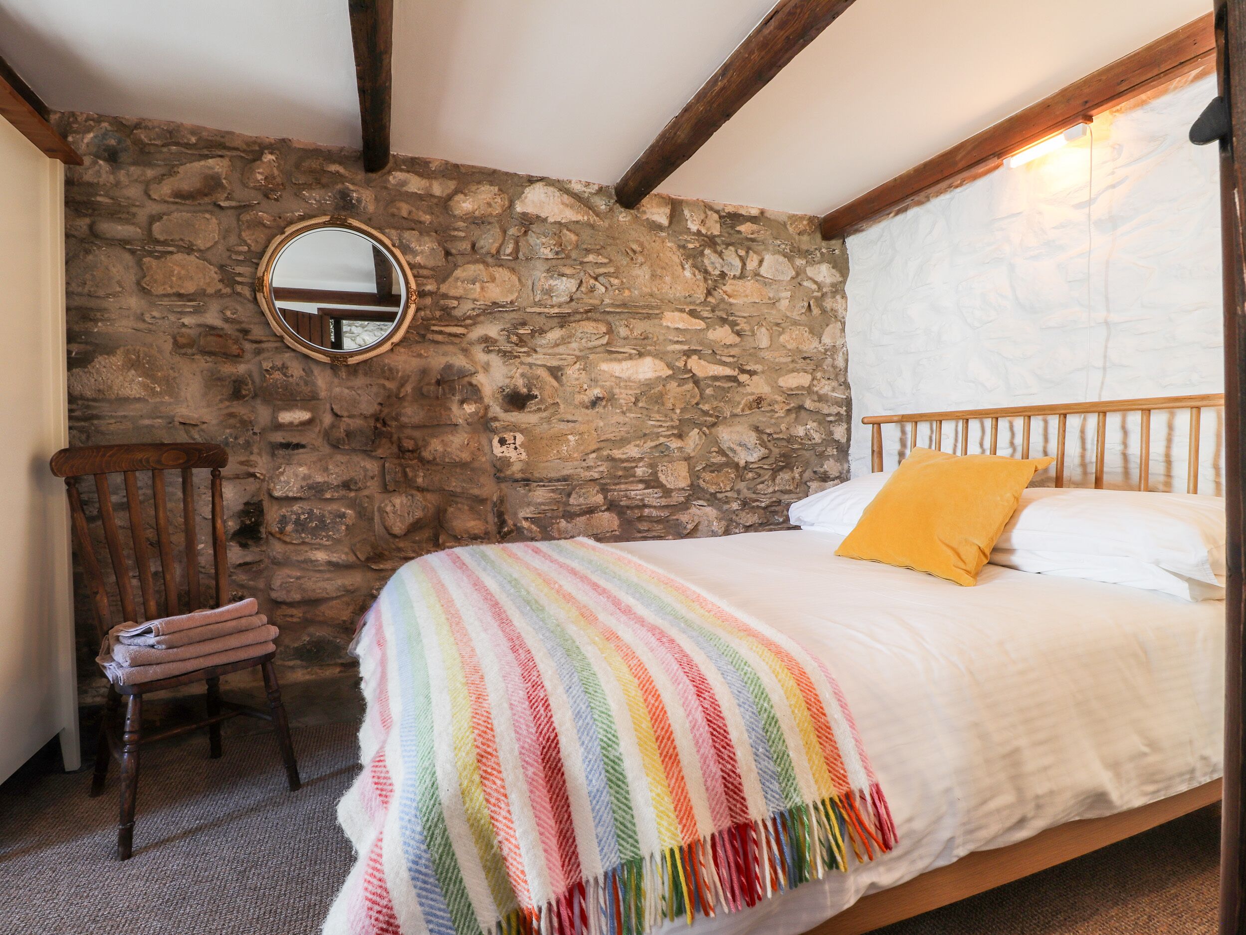 Une chambre avec un mur en pierre, un cadre de lit en bois, une chaise et un miroir rond.