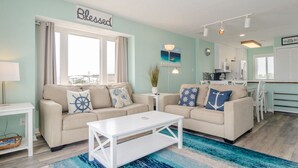 A living room with a sofa, coffee table, and a window with curtains. There is a kitchen area visible in the background. - Top floor ocean front condo in Ocean Dunes! (Kure Beach)