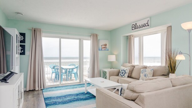 A living room with a sofa, a coffee table, a TV, and a view of the ocean through large windows. - Top floor ocean front condo in Ocean Dunes! (Kure Beach)