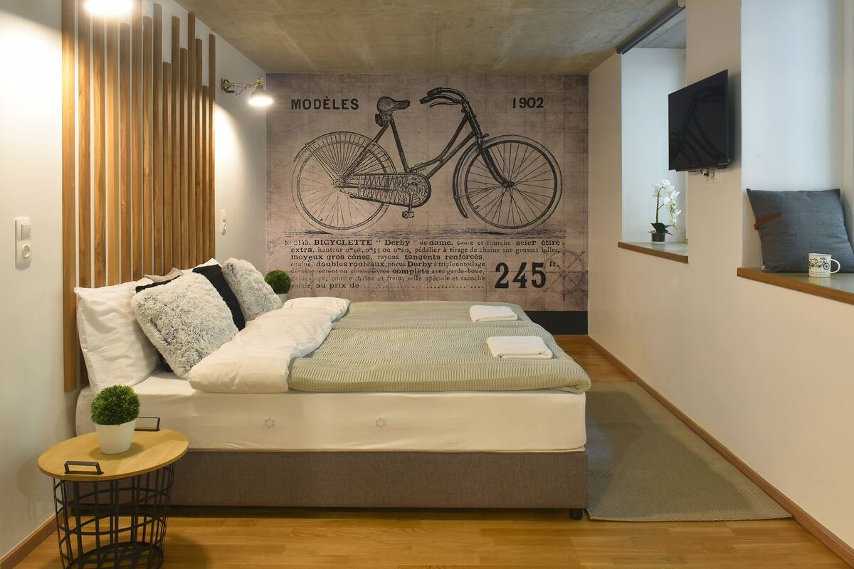 Un dormitorio moderno con cama, una mesita auxiliar, una planta en maceta y una pared con una ilustración de bicicleta y texto.