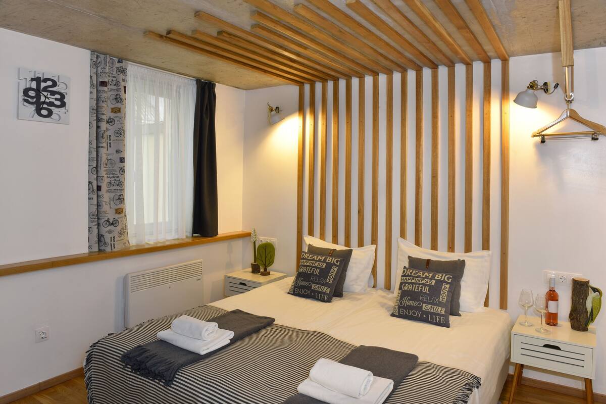 Un dormitorio con cama, almohadas, una mesita de noche, una lámpara colgante y una ventana con cortinas.