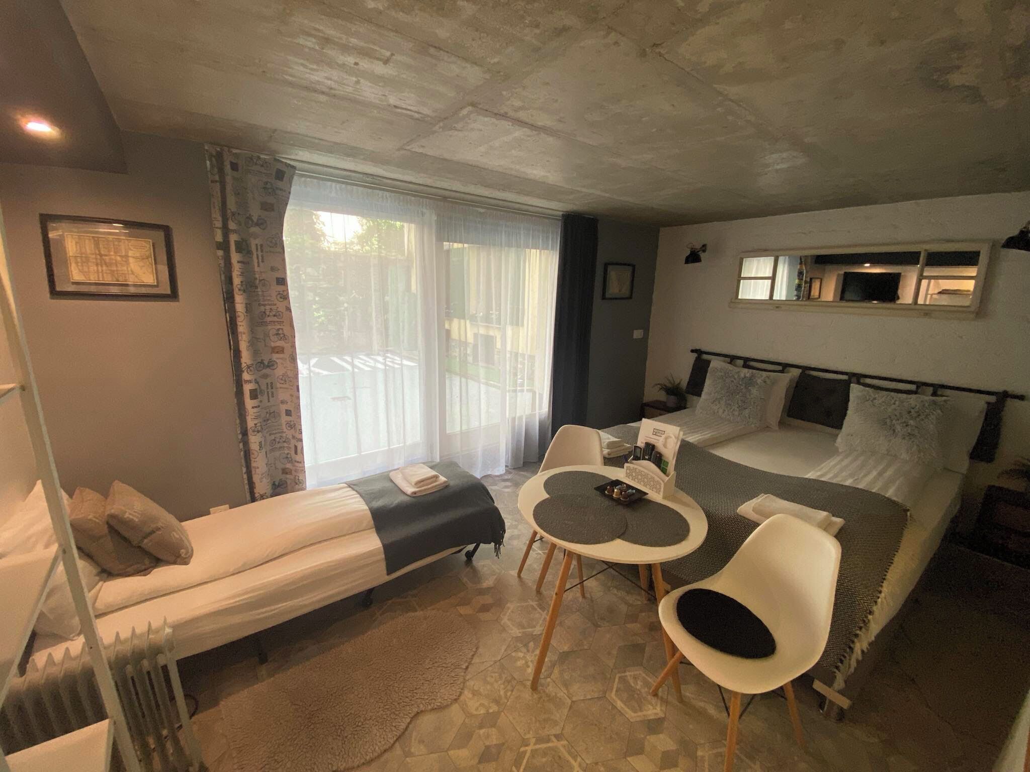 A bedroom with a bed, a small table, two chairs, a radiator, and a window with curtains.