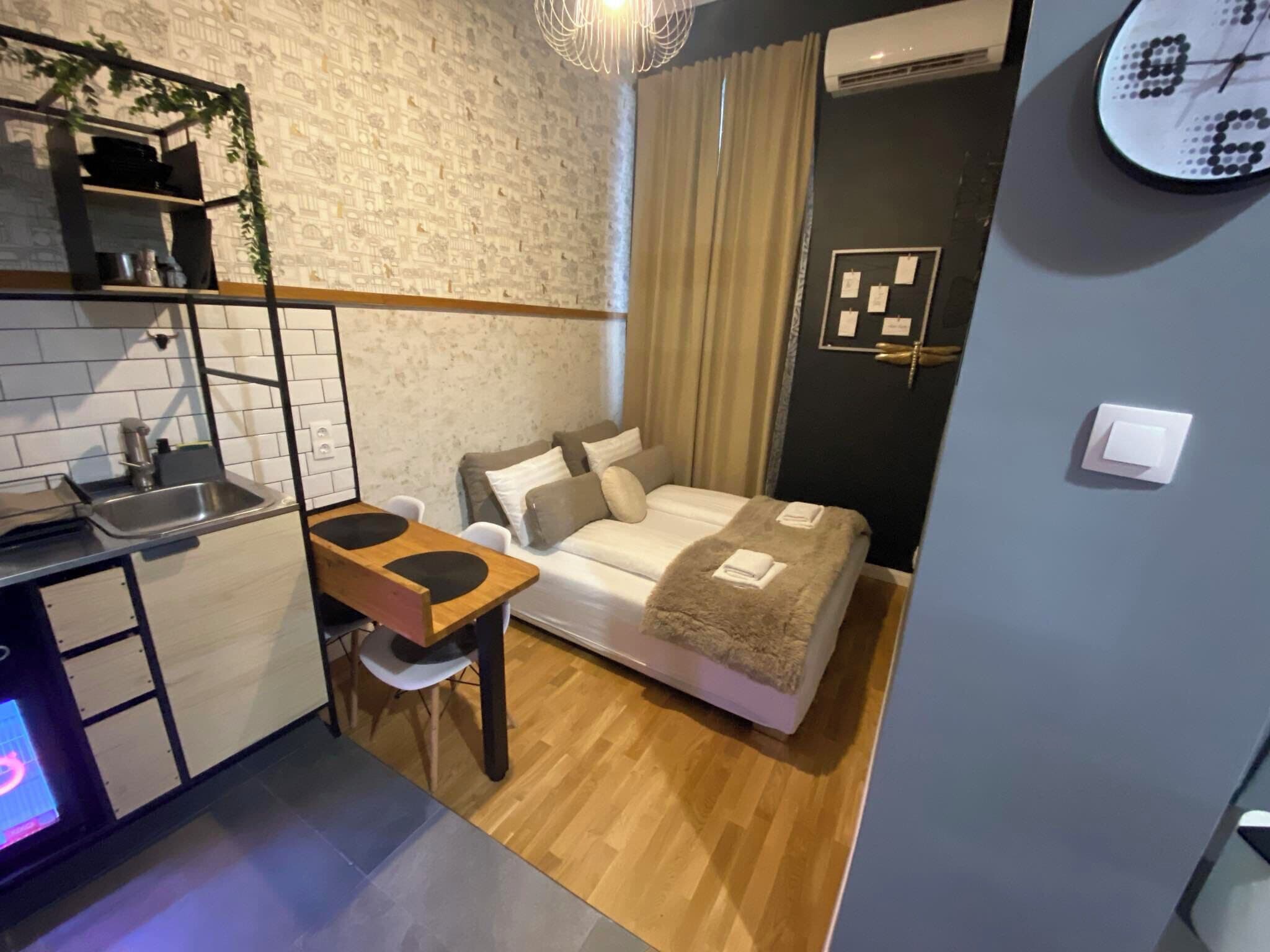 A compact hotel room with a kitchenette, a bed with a fluffy blanket, a small table, and a chair.