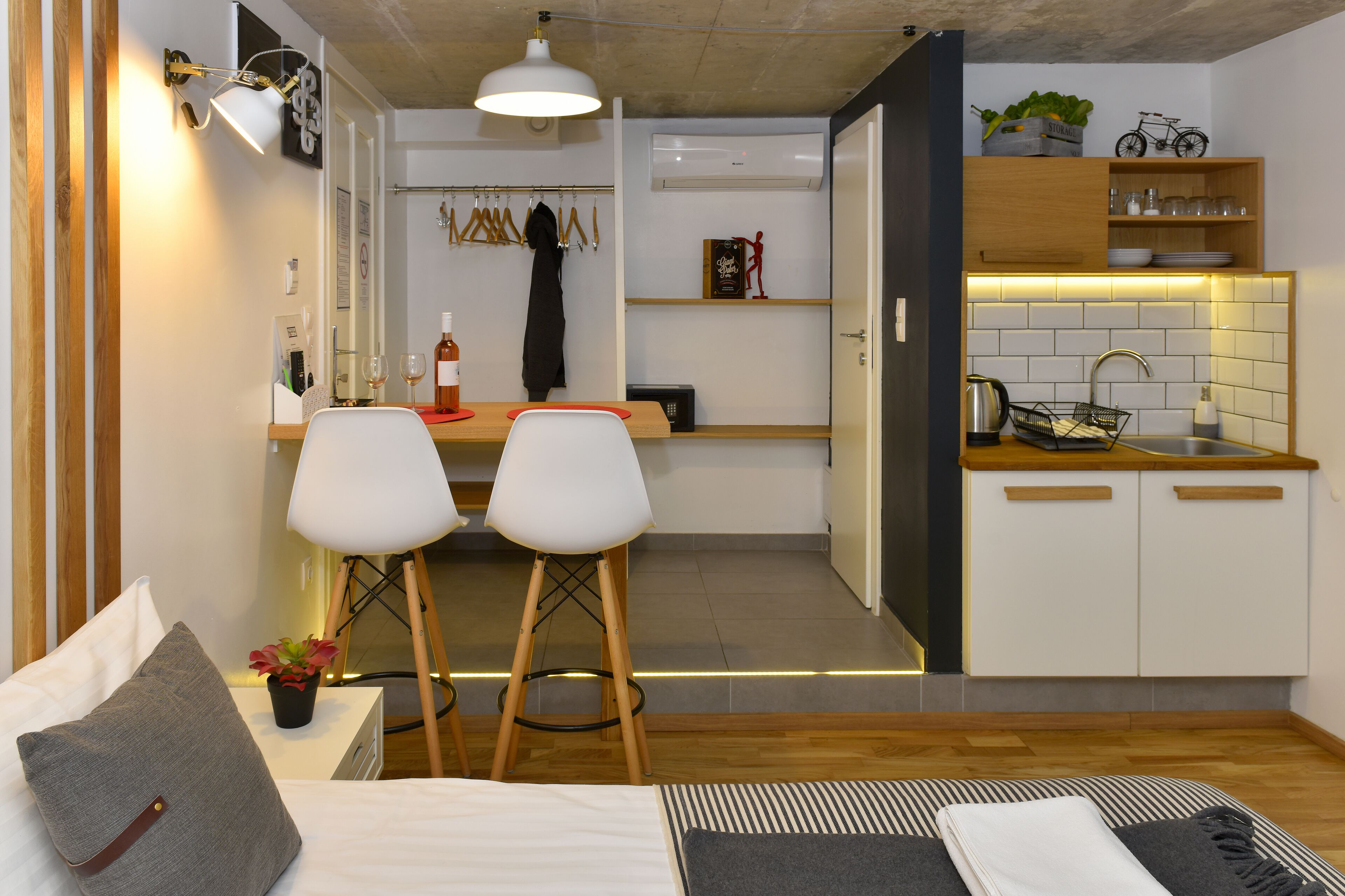 A compact hotel room with a kitchenette, a dining area with two chairs, a bed with a pillow, and a small table with a plant.