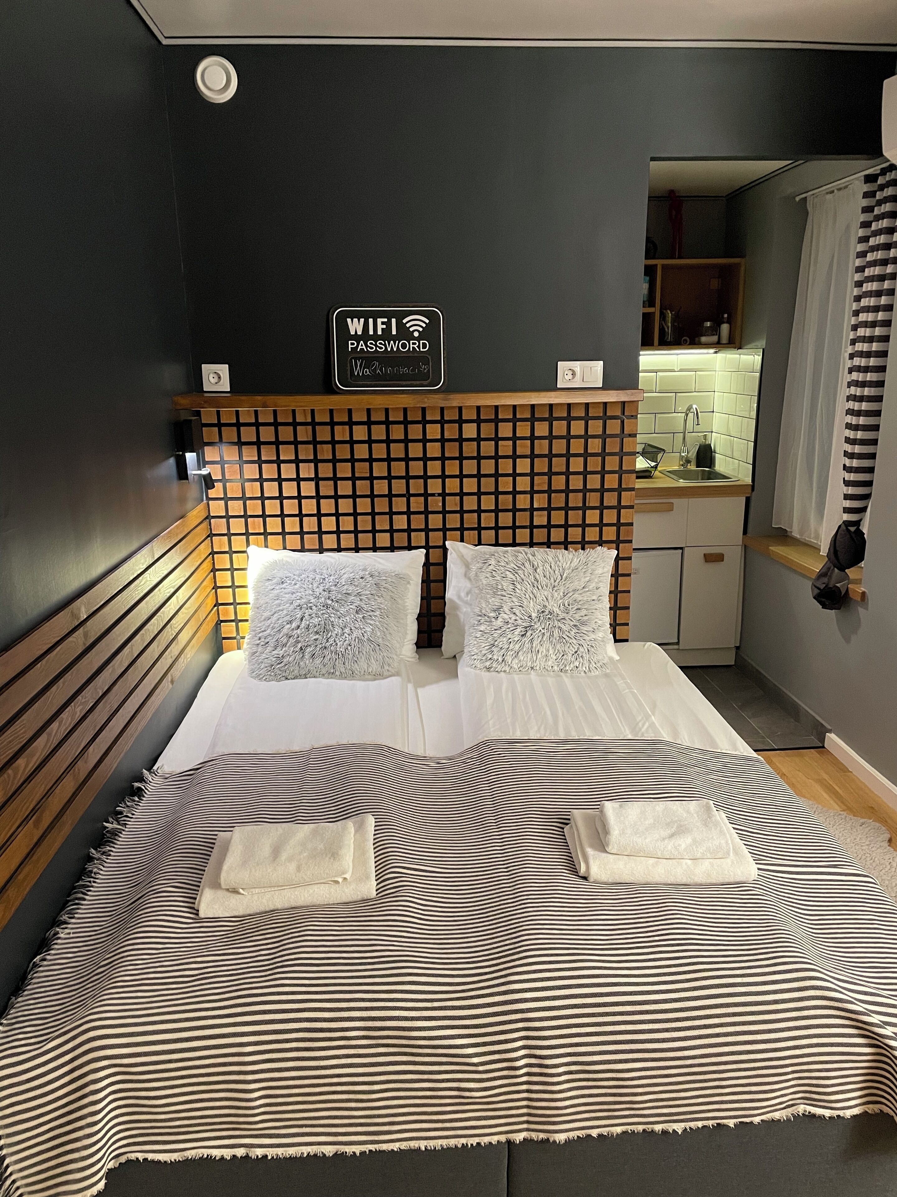 Una camera d'albergo con un letto, una testiera a griglia e un cartello che mostra la password del WiFi, con vista sull'area della cucina.
