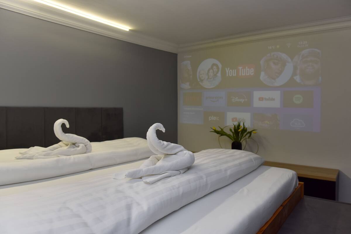 Habitación de hotel con dos camas, cada una adornada con una toalla con forma de cisne. Se observa una pantalla en la pared mostrando YouTube y otras plataformas de video.