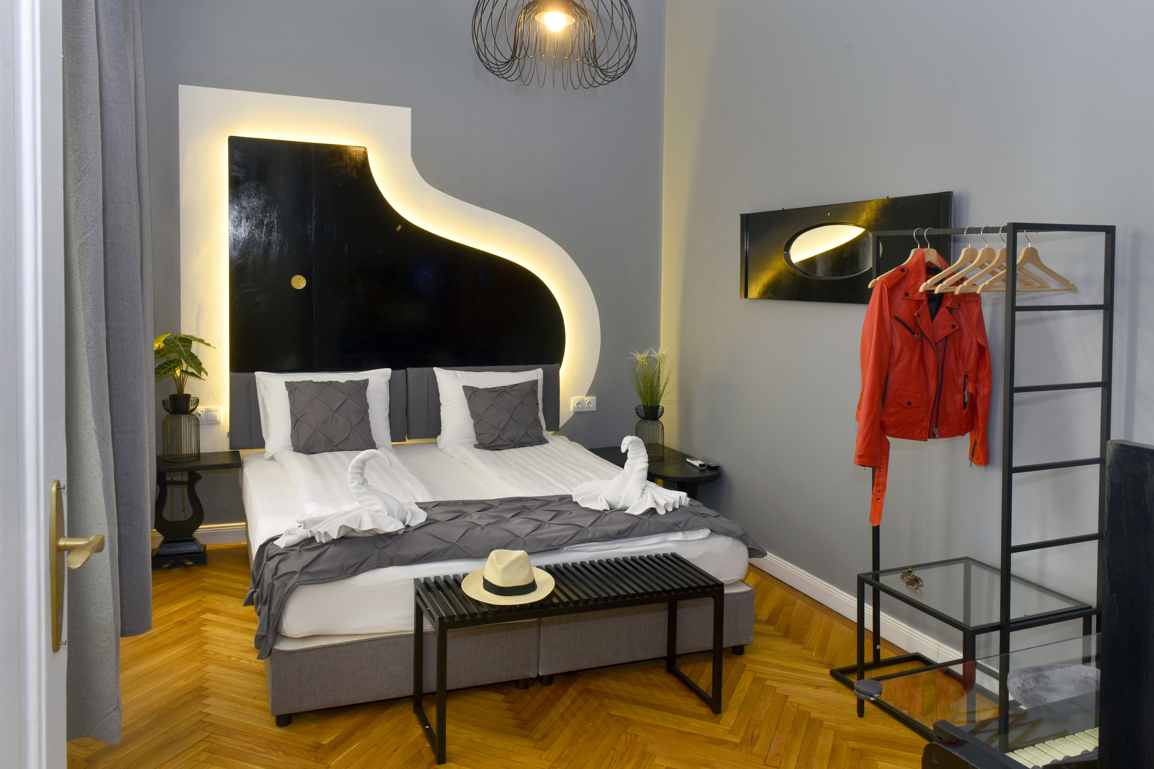 A modern bedroom with a curved headboard, a bed with white linens, a bedside table with a hat, and a coat rack with a red jacket.