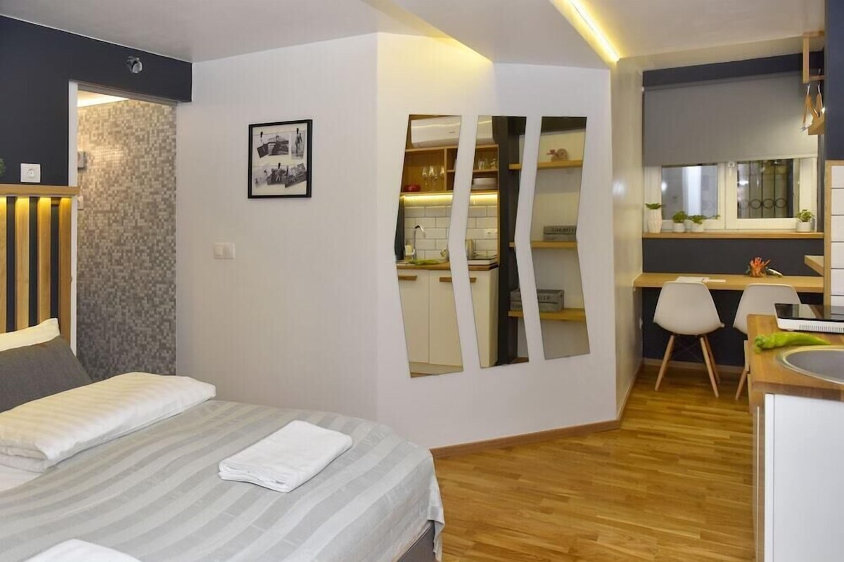 A modern hotel room with a bed, a desk, a chair, and a kitchenette.