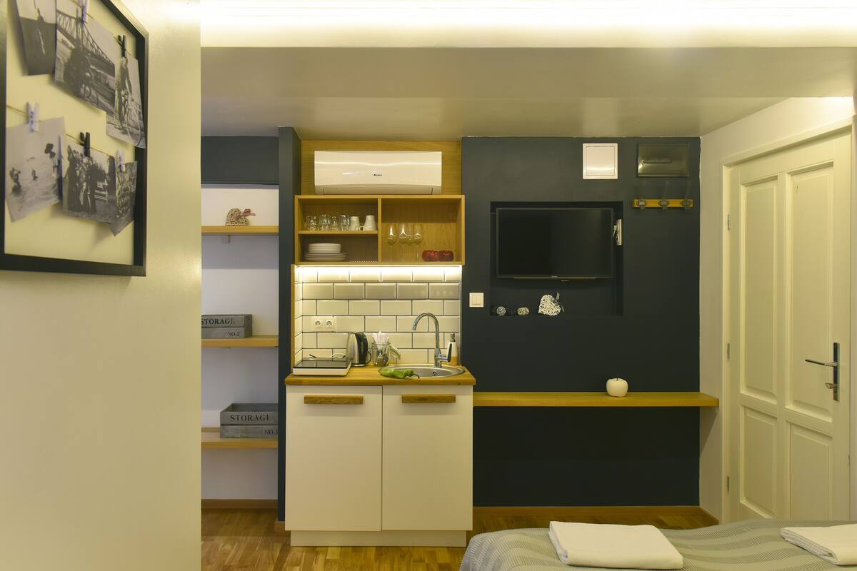 Cocina compacta con lavamanos, microondas y un refrigerador pequeño. Hay repisas de madera sobre el lavamanos y un televisor de pantalla plana montado en la pared.