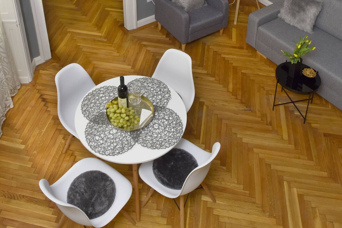 Una sala moderna con piso de madera en patrón chevron, sofás grises, una mesa de comedor con un centro decorativo, y una lámpara colgante de ratán.
