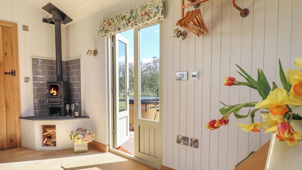 Une pièce chaleureuse avec une cheminée, un plancher en bois, une porte donnant sur l’extérieur et une petite table sur laquelle est posé un vase de fleurs.