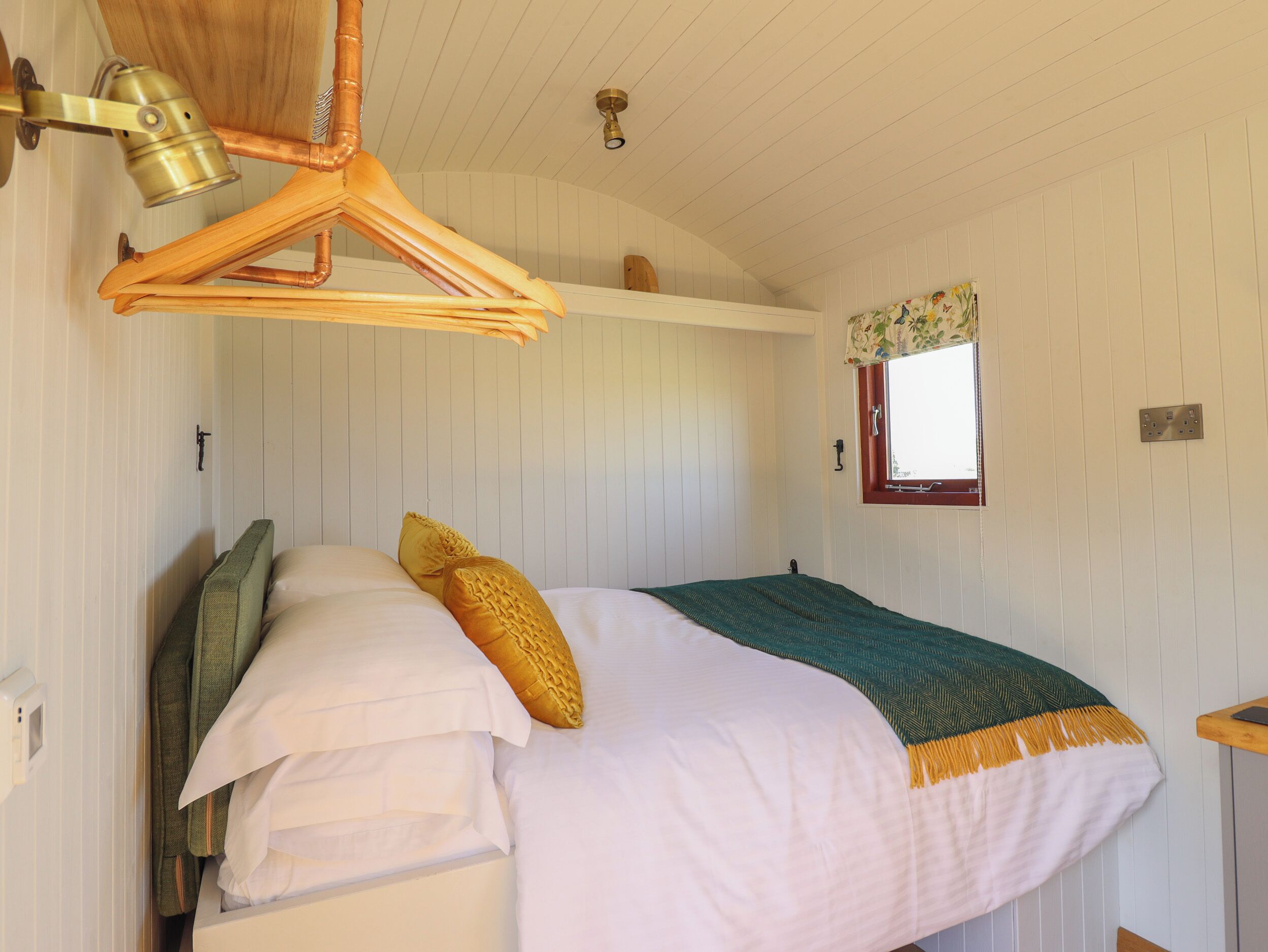 A bedroom with a bed, pillows, a hanging light fixture, a window with a floral curtain, and a wooden ceiling.