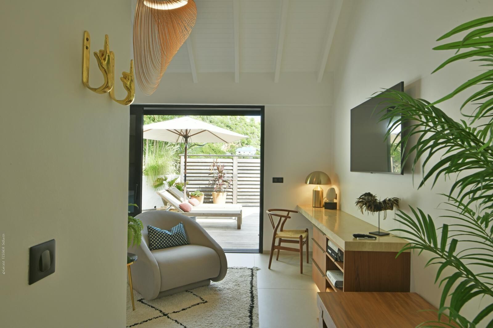 A modern living room with a sofa, a dining area with a table and chairs, a TV mounted on the wall, and a sliding glass door leading to an outdoor patio with plants and a sun lounger.