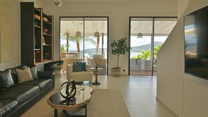 Un salon moderne avec un canapé en cuir, une table basse en verre et une bibliothèque. Une grande porte-fenêtre coulissante donne accès à un balcon aménagé avec du mobilier d’extérieur et offrant une vue sur l’océan.