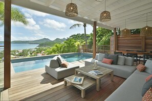 A poolside area with a view of the sea and mountains, wooden deck, outdoor seating, and hanging wicker lamps.