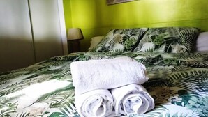 Ett hotellrum med en säng som har ett överkast med ett tropiskt bladmotiv, en prydligt vikt vit handduk på sängen och en tavla med tropiska blad på väggen.