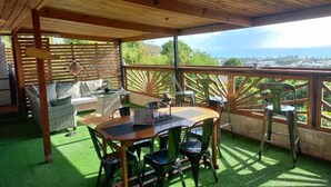 Ett takförsett utomhusområde med sittplatser, bestående av ett träbord och stolar, en soffa och utsikt över havet.