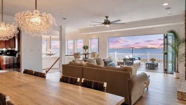 A spacious living area with a large wooden dining table, a brown sofa set, and a view of the sea through large windows.