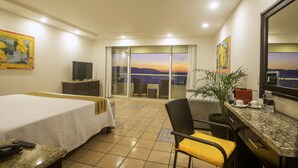 Habitación de hotel con cama, escritorio con silla, televisión, planta y vista al mar.