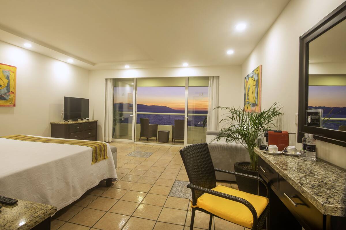 A hotel room with a bed, a desk with a chair, a television, a plant, and a view of the sea.