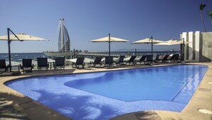 Una piscina con sillas de descanso y sombrillas, un puente y vista al océano.