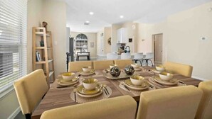 A dining area with a long table set for a meal, a kitchen in the background, and a bookshelf to the left.