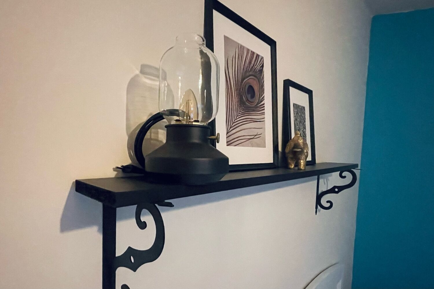 Una estantería de metal negro con un frasco de vidrio, una tetera negra y dos fotografías enmarcadas sobre una pared blanca.