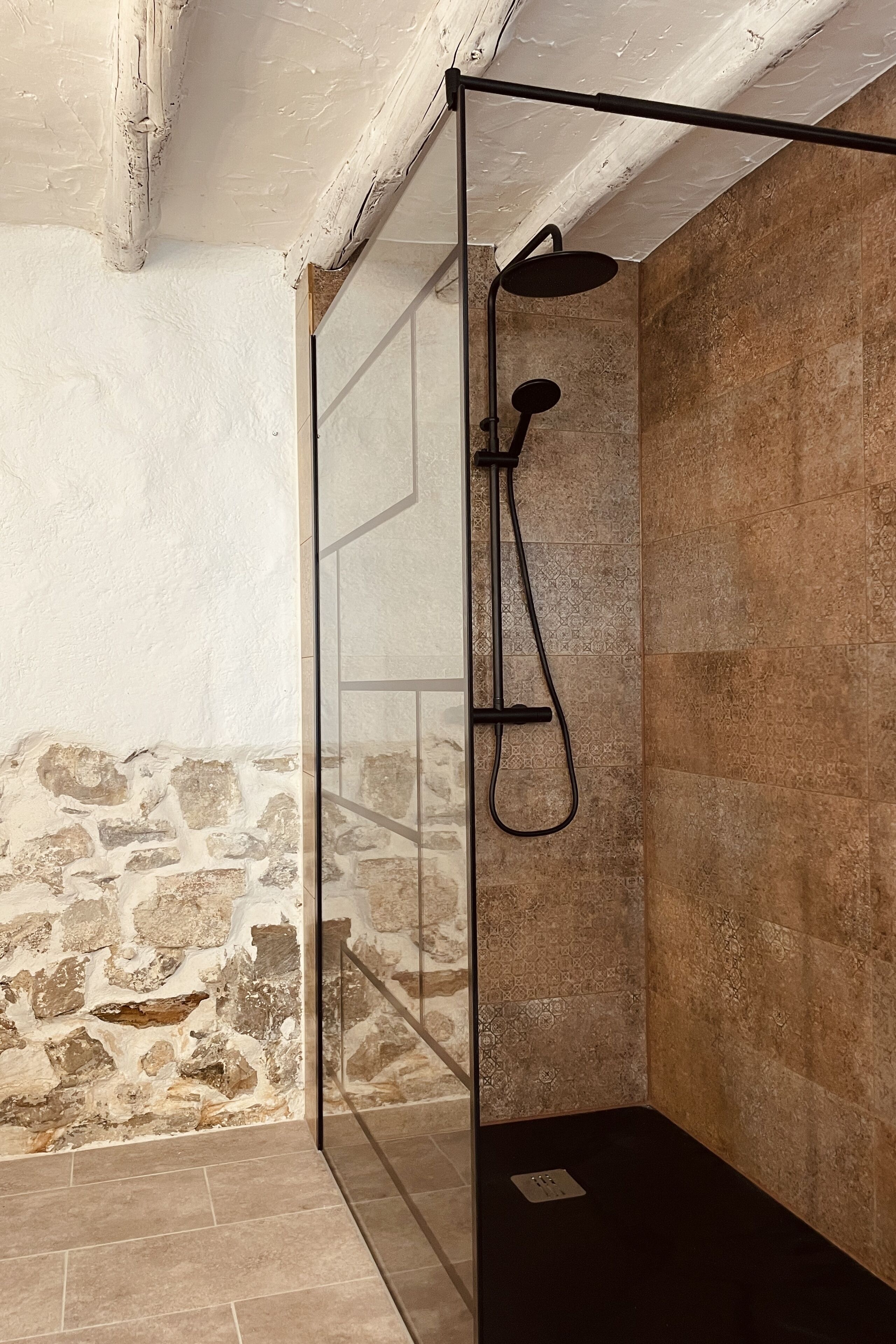 A glass-enclosed shower with a black showerhead and hose, stone walls, and a tiled floor.