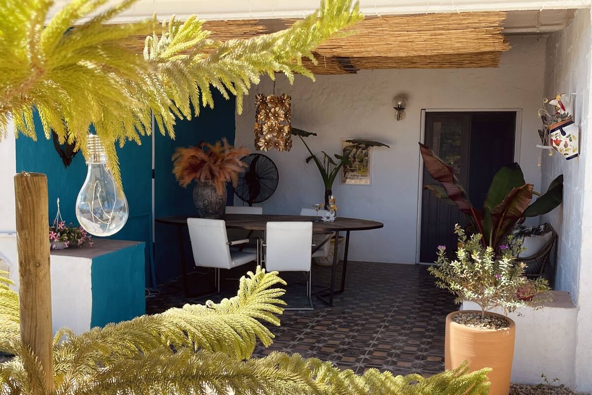 An outdoor dining area with a table and chairs, surrounded by potted plants and a decorative light fixture.