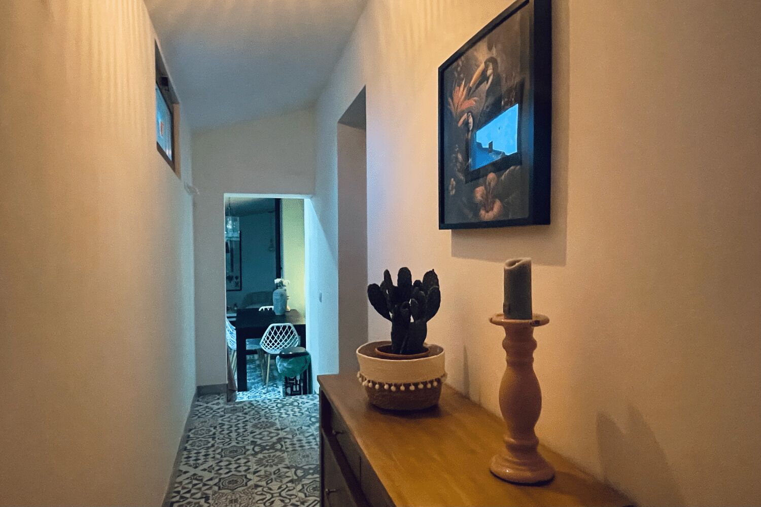 Ein Flur mit einem gemusterten Boden, einem Holztisch mit Kerze und Kaktee sowie einem gerahmten Bild an der Wand.