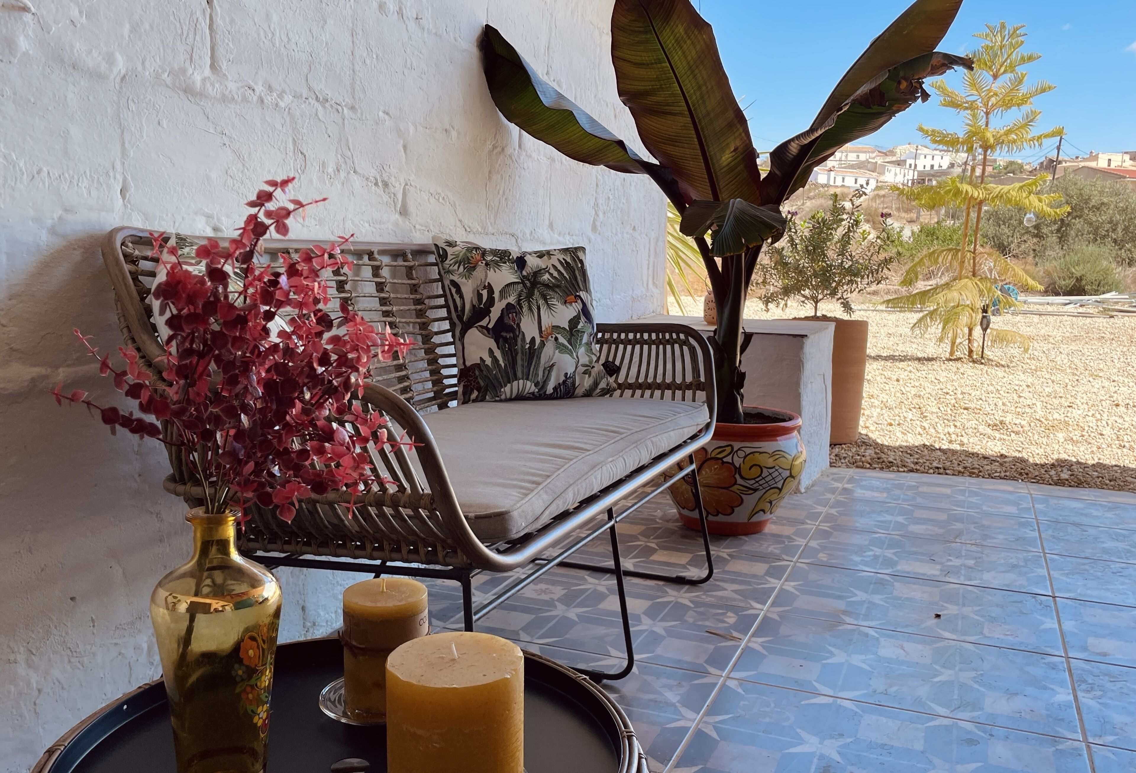Eine Terrasse mit einer Korbsitzgruppe, einem Topfpflanzen, einer Vase mit roten Blättern und zwei Kerzen auf einem Tablett.