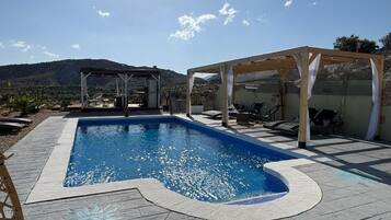 Una piscina con muelle de madera, zona de estar cubierta y montañas al fondo.