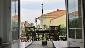 Une porte-fenêtre coulissante donne sur une terrasse extérieure aménagée avec une table et des chaises, un luminaire suspendu et une vue sur un bâtiment jaune doté d’un toit en tuiles.