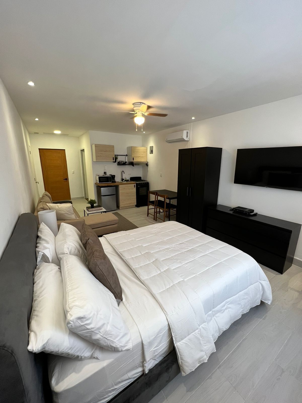A modern hotel room with a large bed, a TV, a kitchenette, and a ceiling fan.