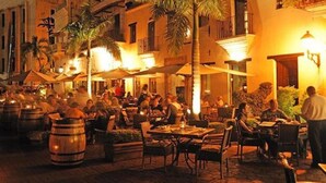 An outdoor dining area with tables and chairs, surrounded by palm trees and buildings. - Suites By Salcedo 3 (Santo Domingo)