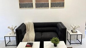 Un salon avec un canapé noir, deux tables blanches à structure noire, un tapis beige et trois œuvres d’art abstraites accrochées au mur.