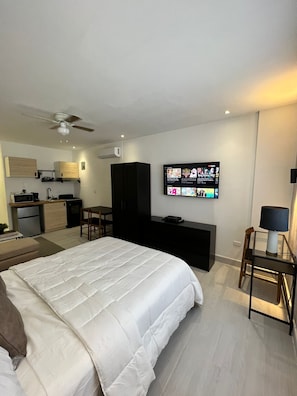 A modern hotel room with a bed, a TV mounted on the wall, a desk, and a kitchenette in the background.