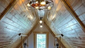 Une chambre mansardée chaleureuse, avec un plafond en pente en bois, un lit recouvert d’une couverture colorée, deux tables de chevet et une fenêtre laissant entrer la lumière naturelle.