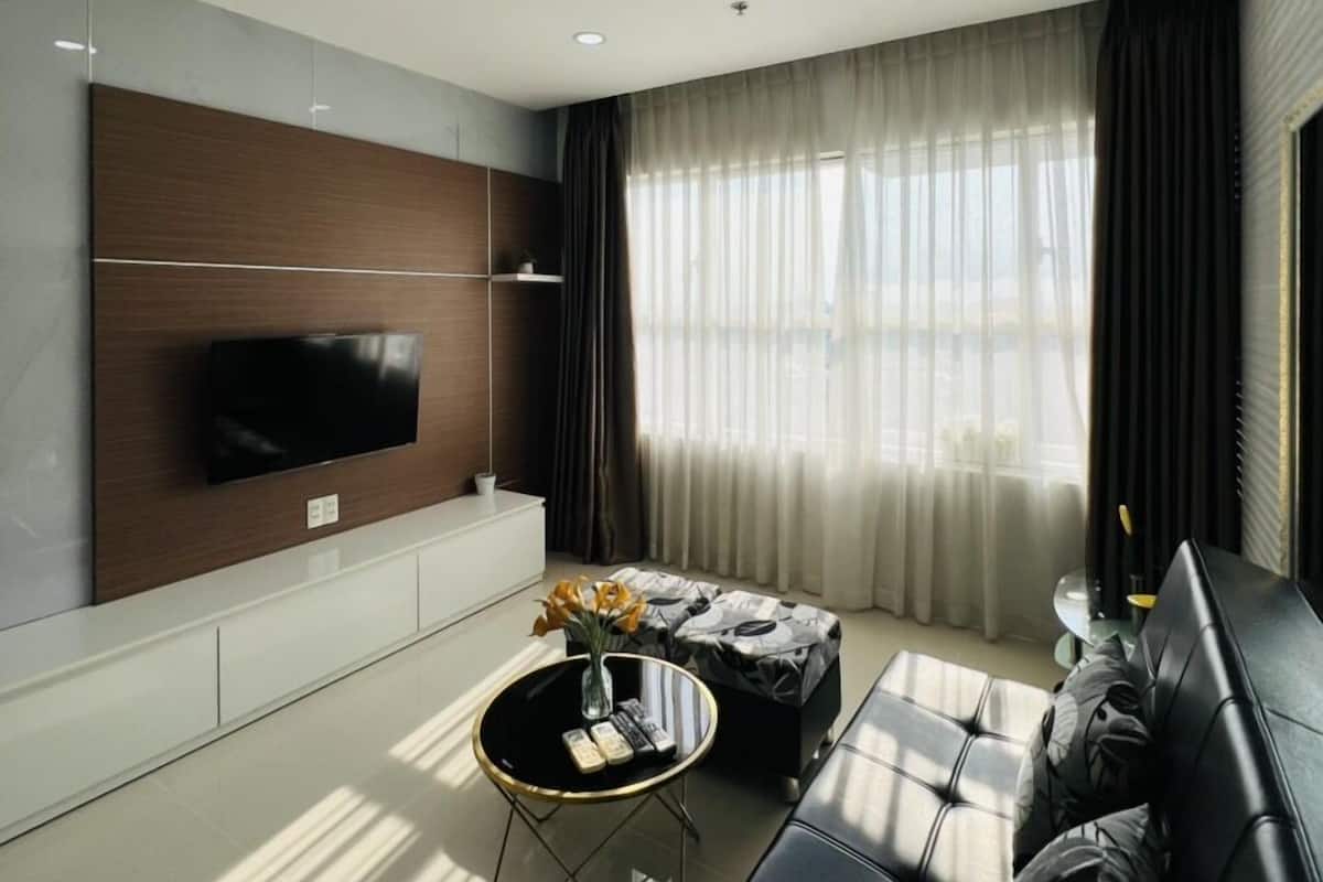 Ein modernes Wohnzimmer mit einem an der Wand befestigten Flachbildfernseher, einer schwarzen Ledersofa, einem Couchtisch und einem großen Fenster mit durchsichtigen Vorhängen.