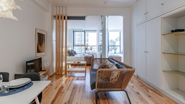 Un soggiorno moderno con pavimento in legno, un divano in vimini, un tavolo da pranzo con piatti e bicchieri, e vista su un balcone con un letto e un divano.
