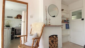 Un fauteuil à bascule en bois avec un coussin moelleux, une cheminée avec des bûches, un miroir et une cuisine avec des meubles blancs et un poêle.