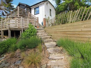 A wooden staircase leading to a house with a balcony, surrounded by greenery and a wooden fence.