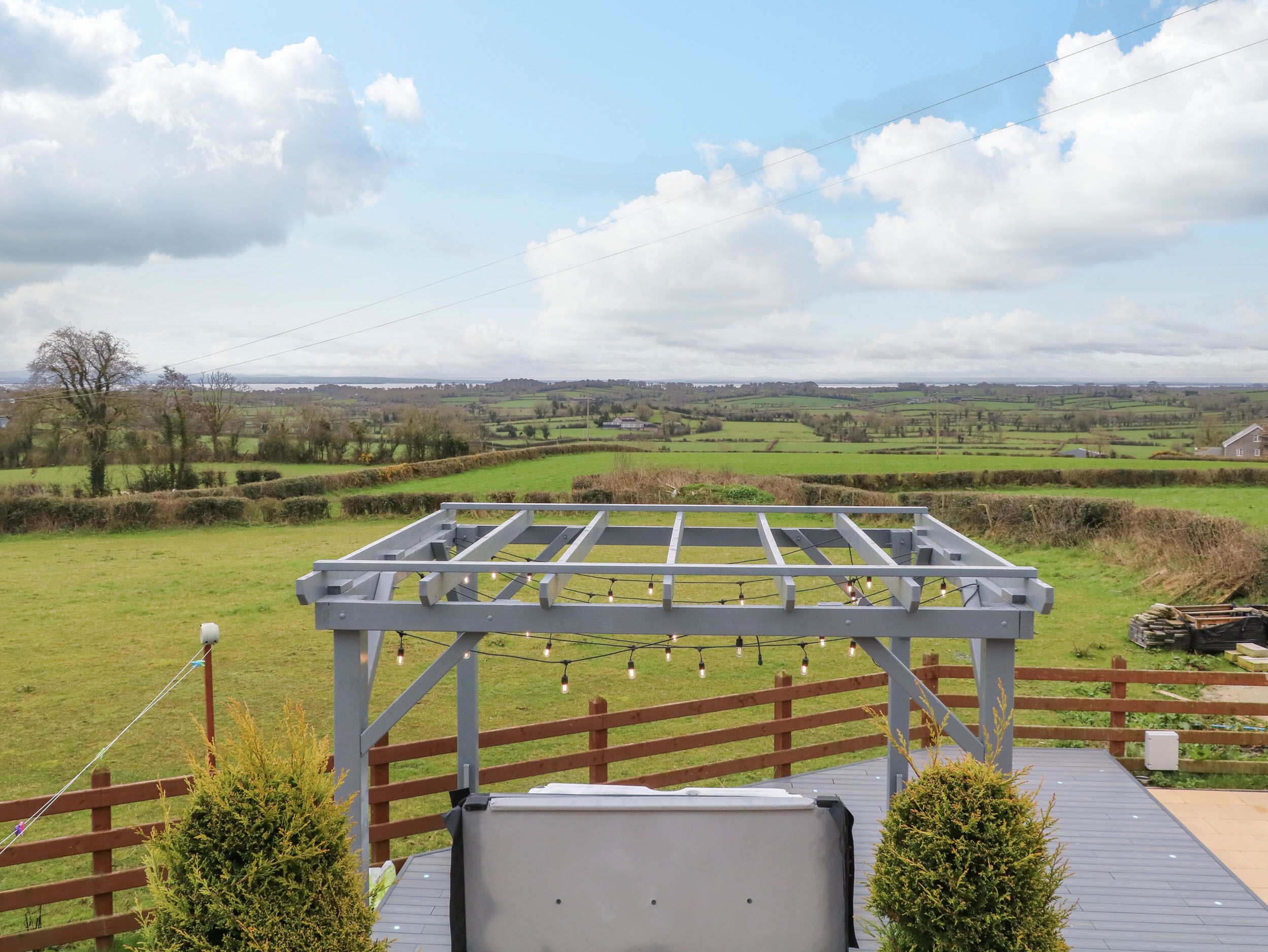 Une terrasse en bois surmontée d’une pergola, offrant une vue sur un paysage rural avec des champs et des bâtiments situés au loin.