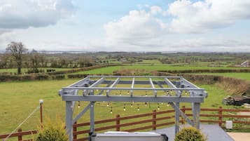 A wooden deck with a pergola structure, overlooking a rural landscape with fields and distant buildings.
