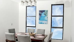 Un comedor con una mesa de madera, sillas tapizadas en gris y una araña. Hay dos ventanas grandes con marcos negros y un cuadro de un paisaje azul.